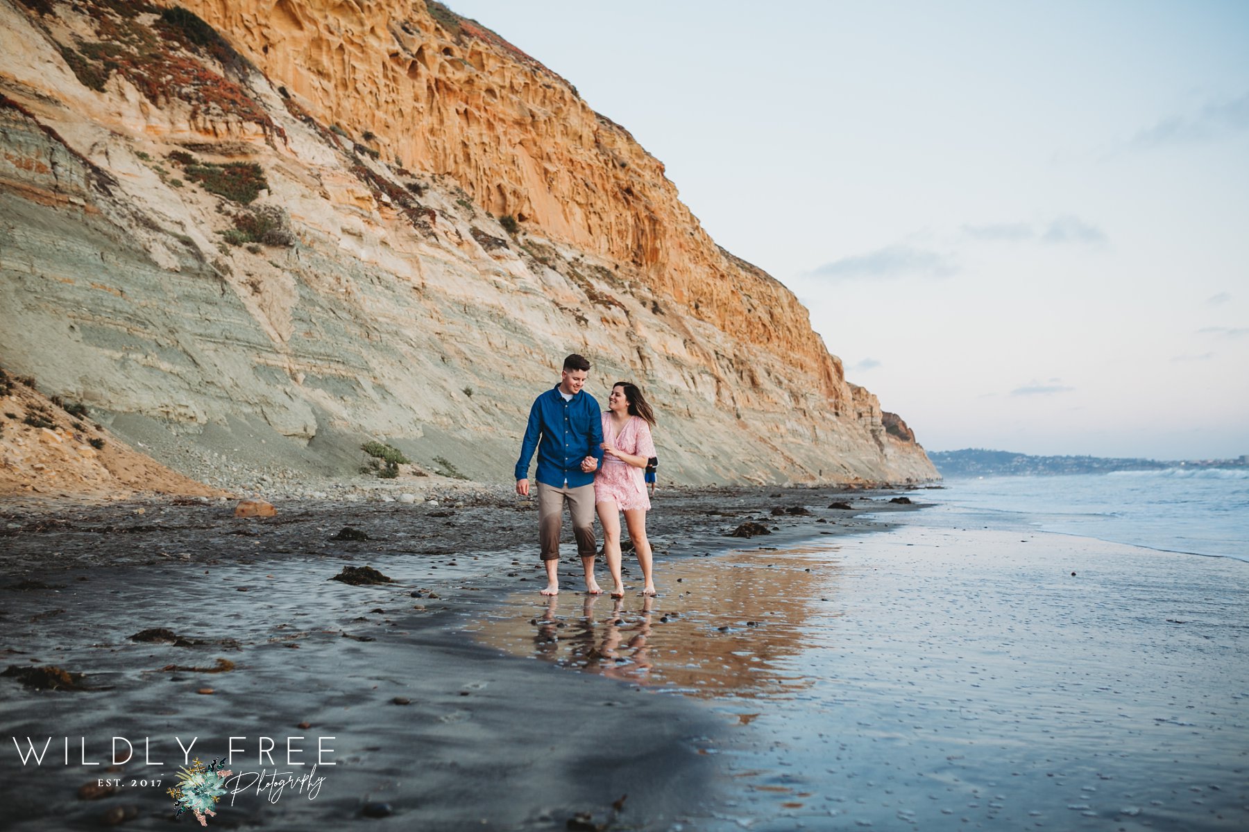 Engaged couple walking on the beach in front of cliff in Torrey Pines CA